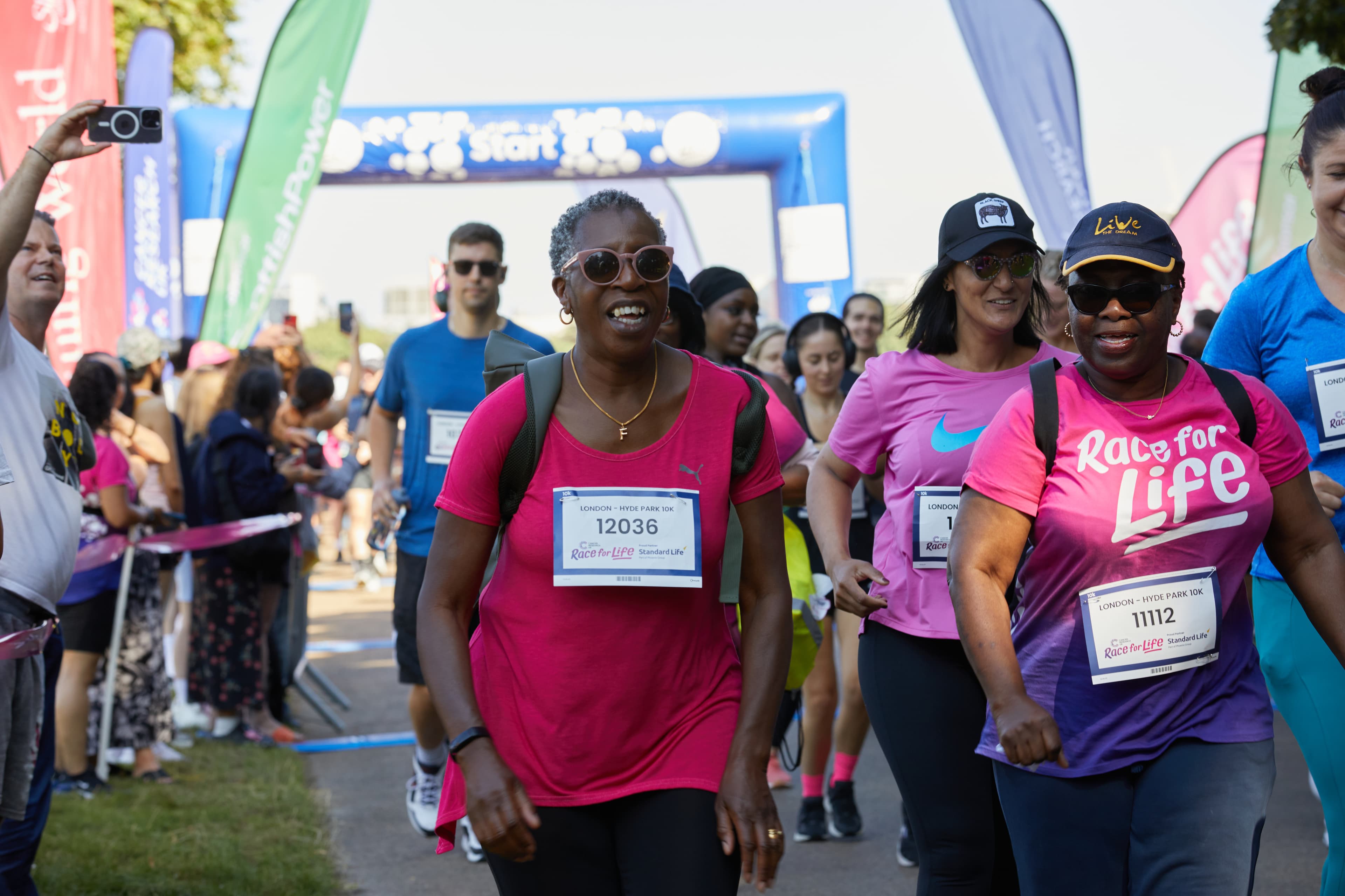 A large group with a pair of women at the front walking through the start line with a crowd on the side.