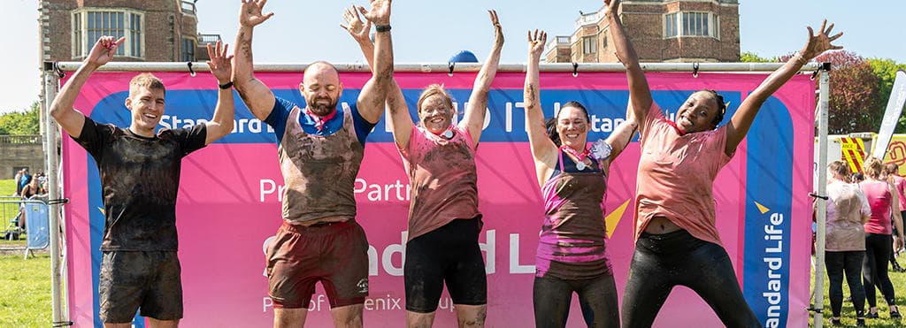 Group of people covered in mud, smiling and jump in front of Proud Partner sign.