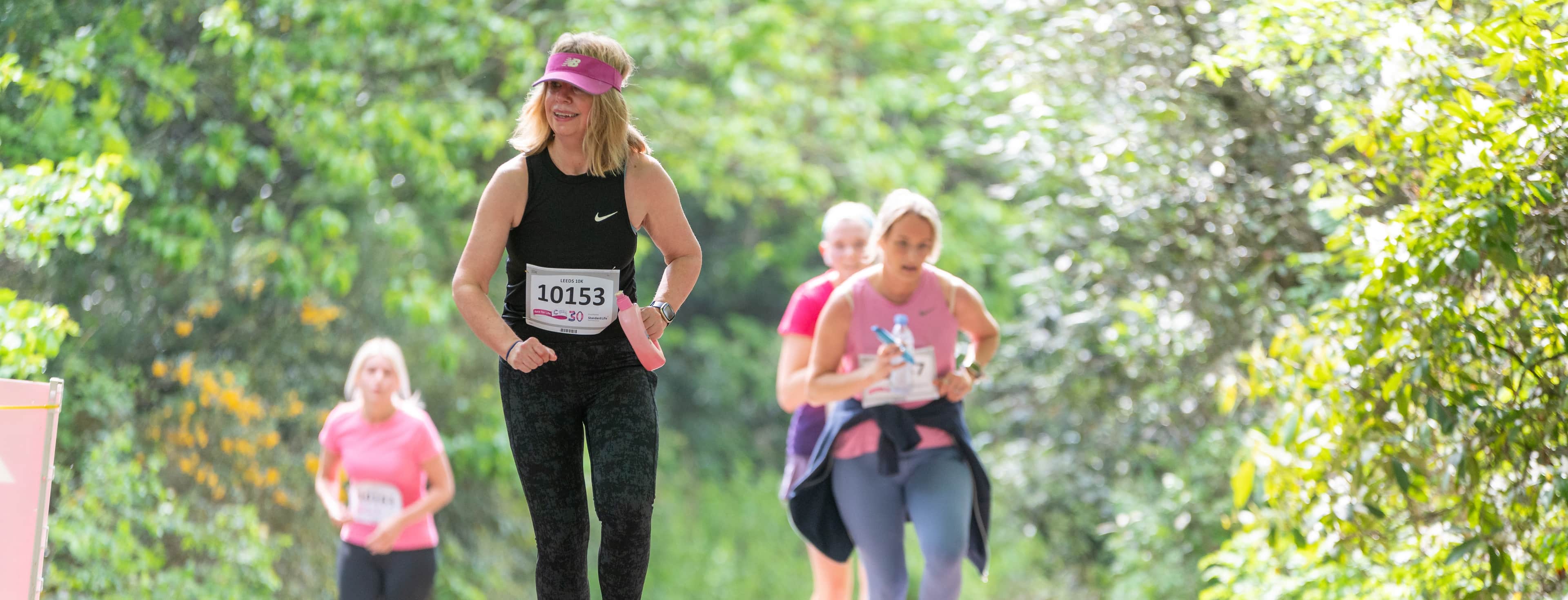 A group of women running in Race for Life t-shirts in a park with trees in the background.