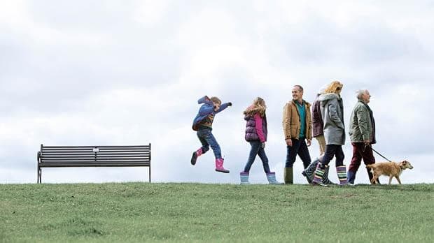 A family of six walking in a park with a small dog past a lone park bench. The youngest is jumping in the air.