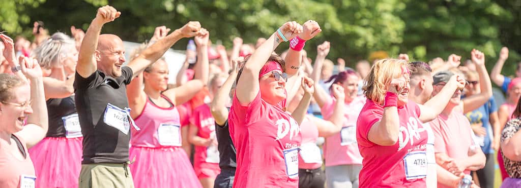 A crowd of Race for Life participants are smiling with their hands in the air. They're warming up before the race begins.